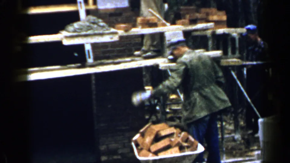 A construction worker moves beside a wheelbarrow of bricks below scaffold planks during 1957 Birmingham house construction.