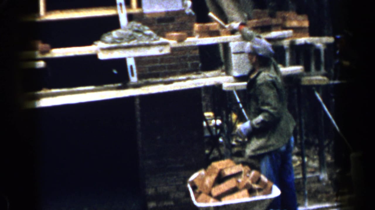 Worker in a cap stands near scaffold planks and a wheelbarrow filled with bricks.