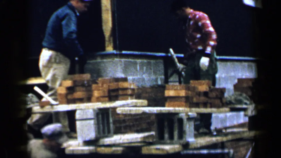Two adult workers in casual clothes handle bricklaying materials inside a Birmingham construction site in 1957.