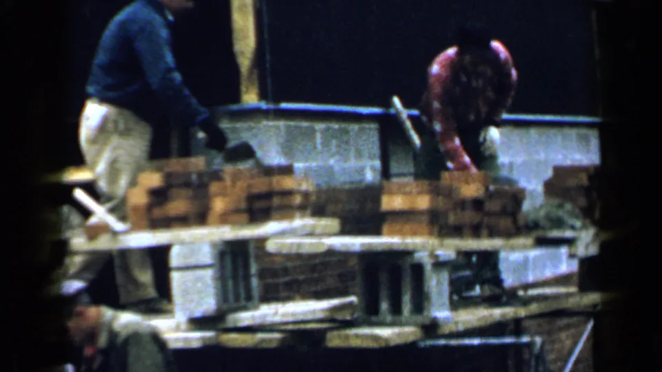 Workers handle bricks and mortar on a partially built wall inside a Birmingham structure, captured on 8mm film in 1957.
