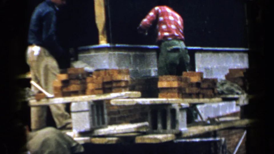 1957 Birmingham construction view with two workers, a red-shirted bricklayer, brick stacks, and concrete blocks on a scaffold.