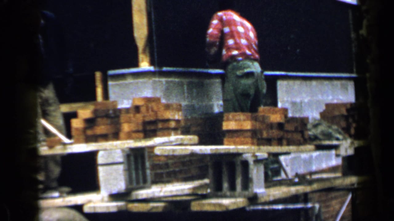 Adult men work around stacked bricks and concrete blocks at a partially built foundation.