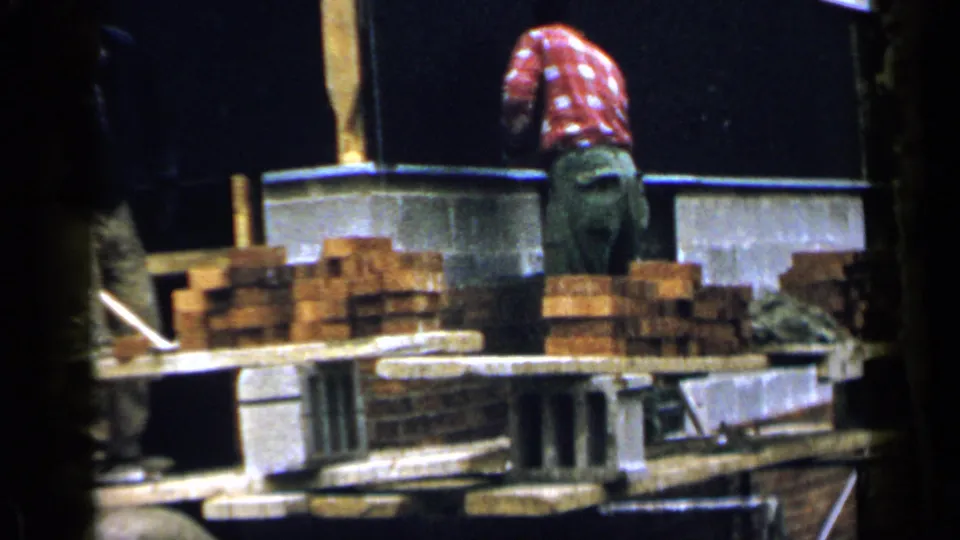 1957 Birmingham home-movie frame showing adult men near stacked red bricks, concrete blocks, and a partially built masonry foundation.