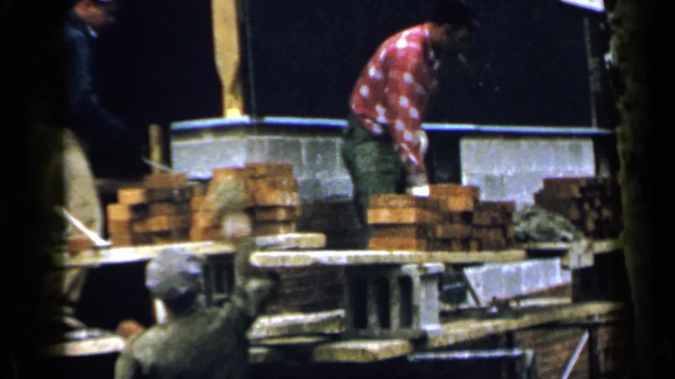 1957 Birmingham frame with a worker in a red checkered shirt handling bricks near concrete blocks and construction planks.