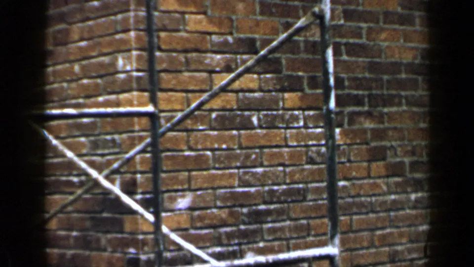 1957 home-movie frame of brickwork, scaffold bracing, and dark 8mm gate edges in Birmingham.