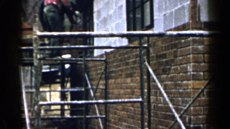 A worker is partly visible above scaffold rails near brick and concrete walls in the 1957 Birmingham construction footage.