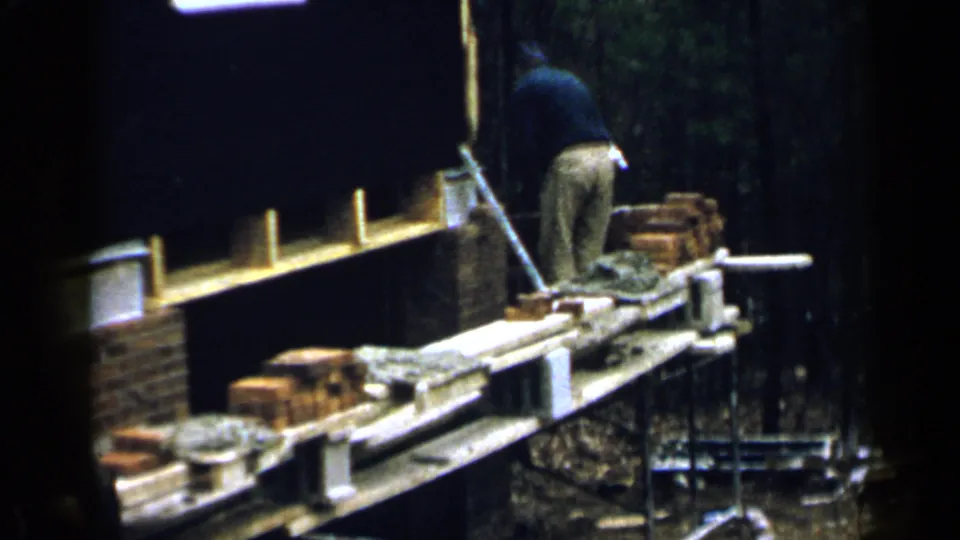 A man bends over a scaffold platform with stacked bricks and mortar during 1957 Birmingham building work.