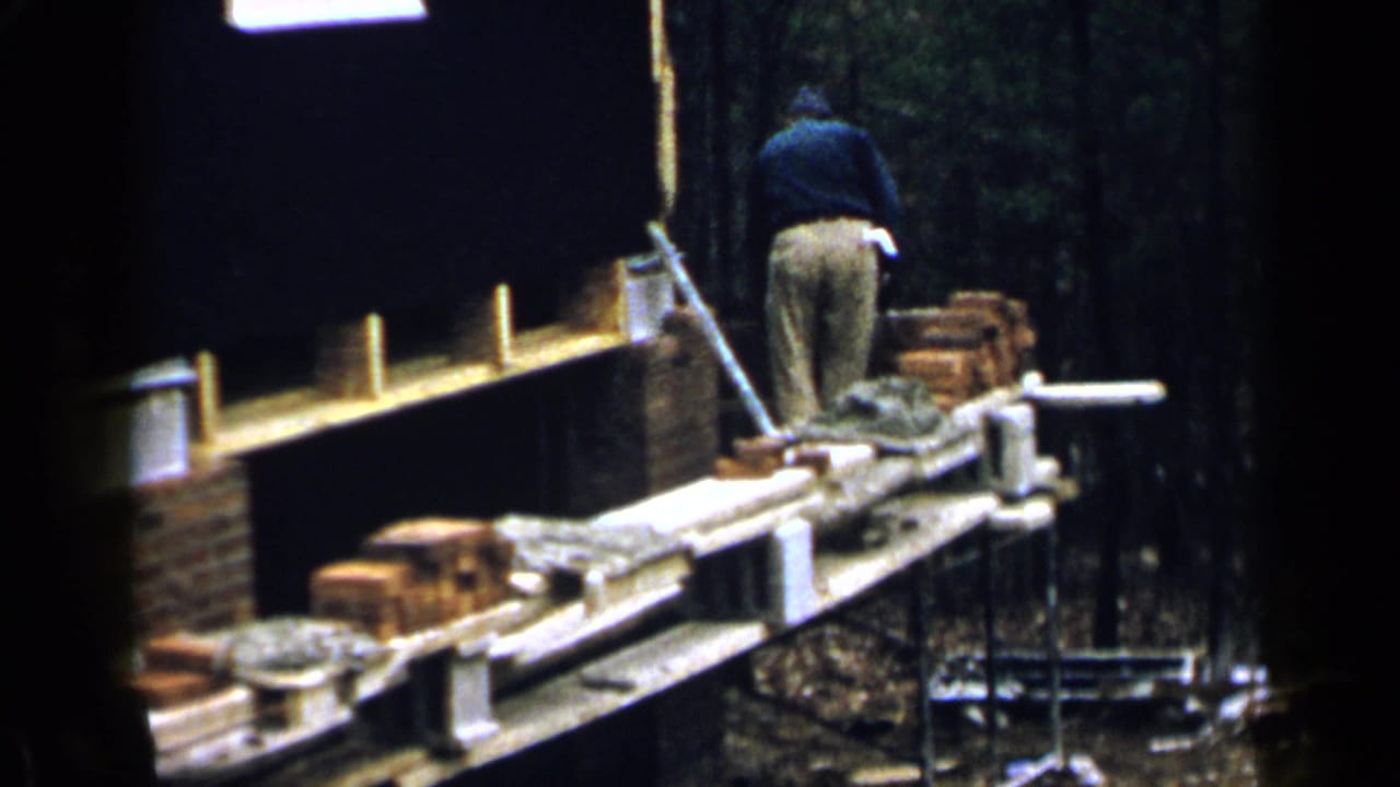 Man working beside a brick wall from a wooden scaffold in a wooded outdoor setting.