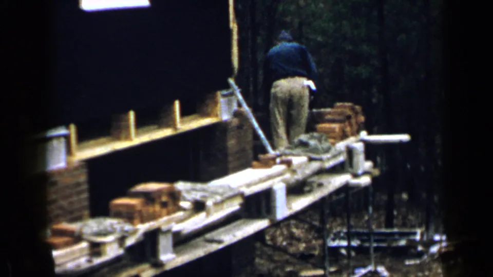 1957 Birmingham 8mm frame showing a man in a dark sweater working on a brick wall from a wooden scaffold outdoors.