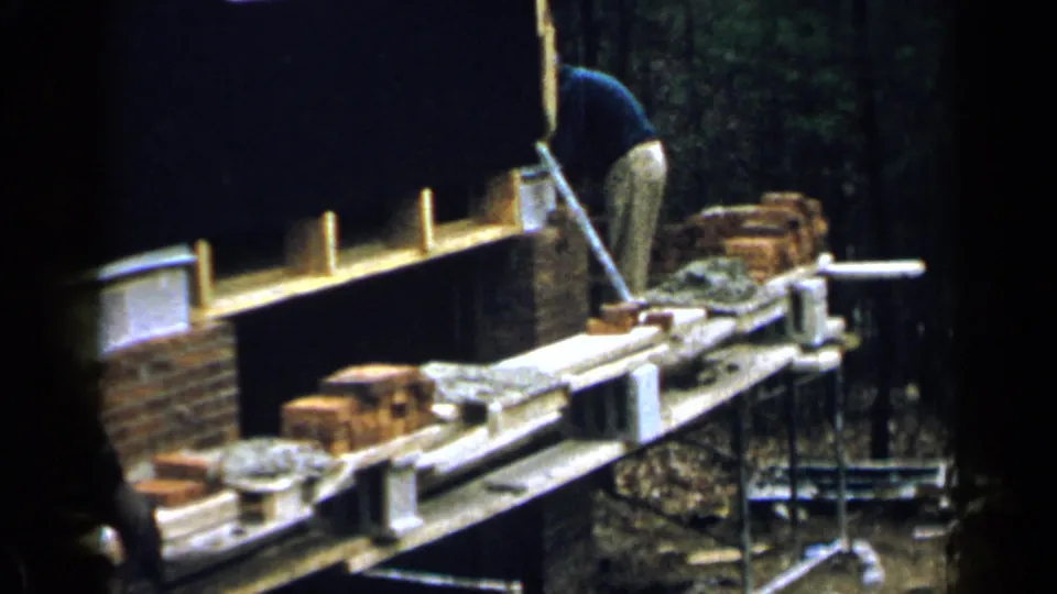 1957 Birmingham home-movie frame of a man leaning over brickwork on a scaffold at an outdoor masonry site.