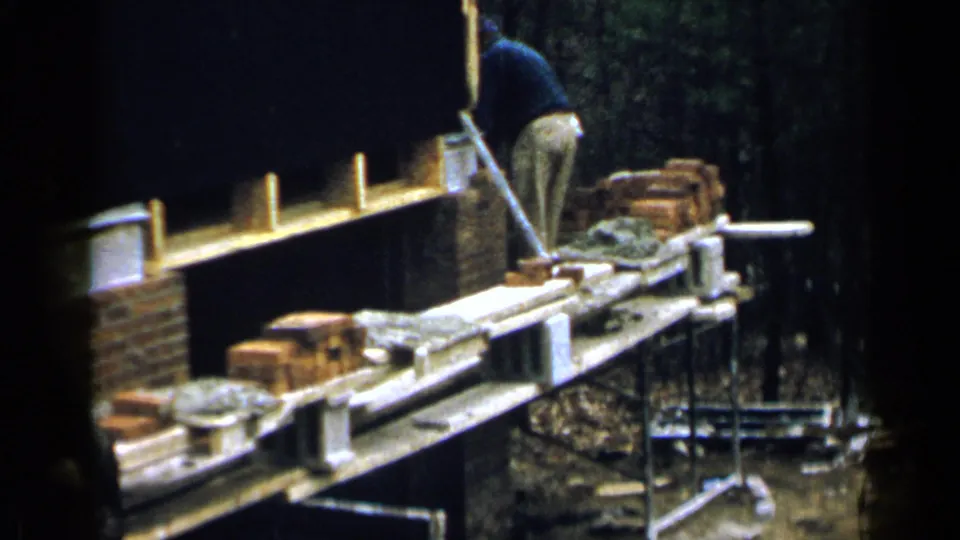 1957 Birmingham 8mm frame showing a man at the far side of a scaffolded brick wall in a wooded outdoor construction area.