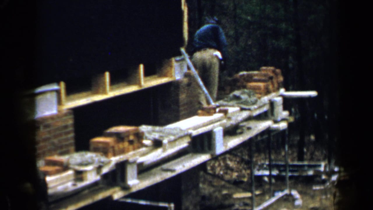 Worker laying bricks along a raised plank at a wooded construction site.