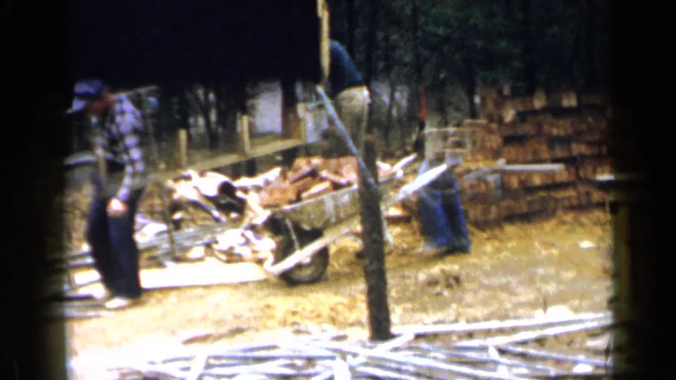 1957 Birmingham-area home-movie frame with a worker moving through a wooded construction site near a wheelbarrow, bricks, and scattered boards.