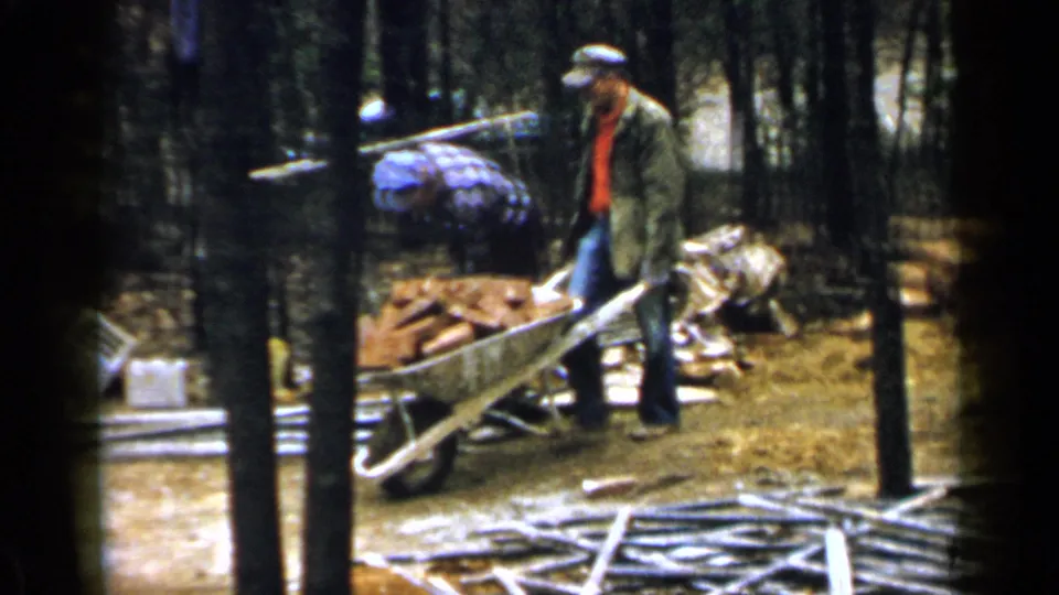 1957 Birmingham-area 8mm frame showing two adult workers around a brick-filled wheelbarrow beside trees and foundation materials.