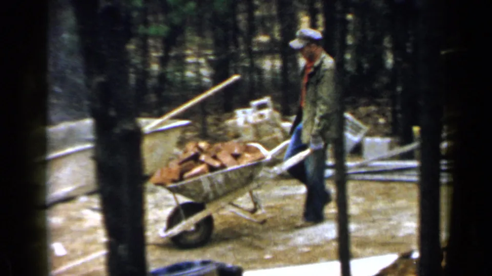 1957 Birmingham-area frame of a worker pushing a brick-filled wheelbarrow through a wooded house foundation site.