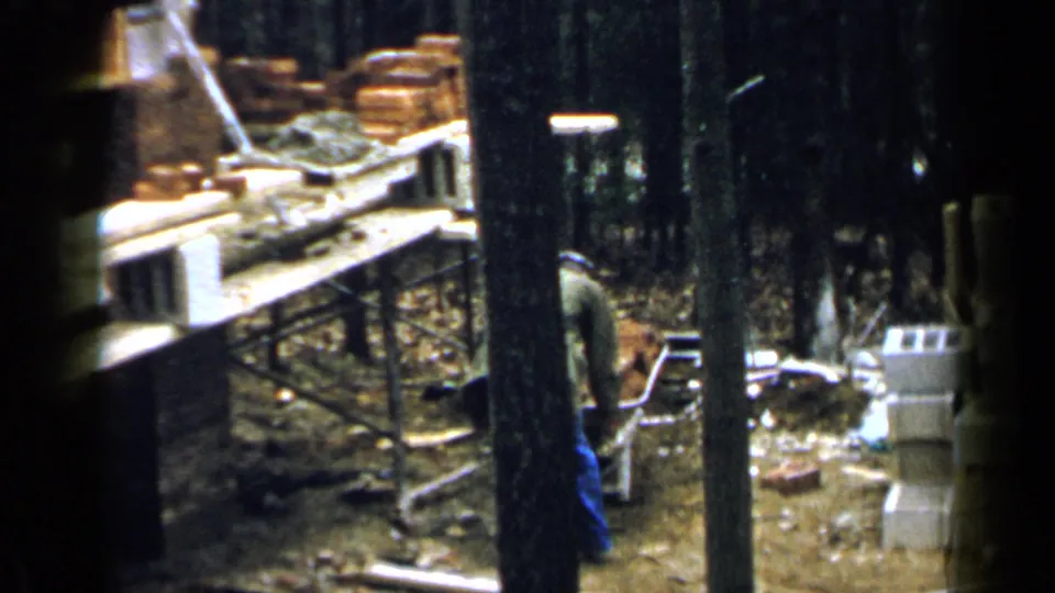 The worker bends near the wheelbarrow as bricks, cinder blocks, planks, and trees fill the 1957 Birmingham work area.