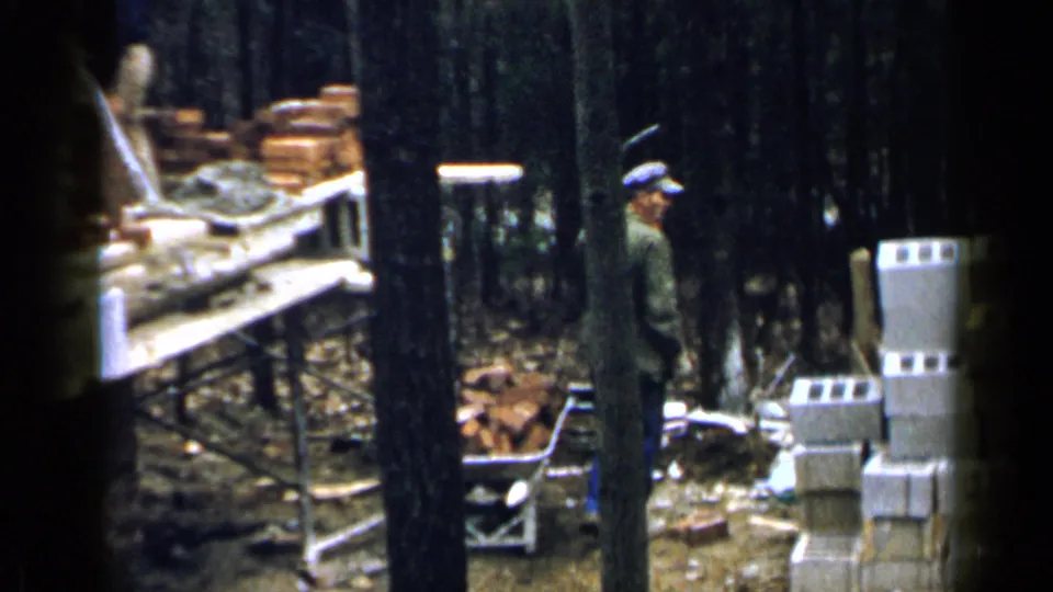 At the end of the clip, the Birmingham worker stands near the wheelbarrow amid stacked blocks, loose bricks, and wooded construction materials.