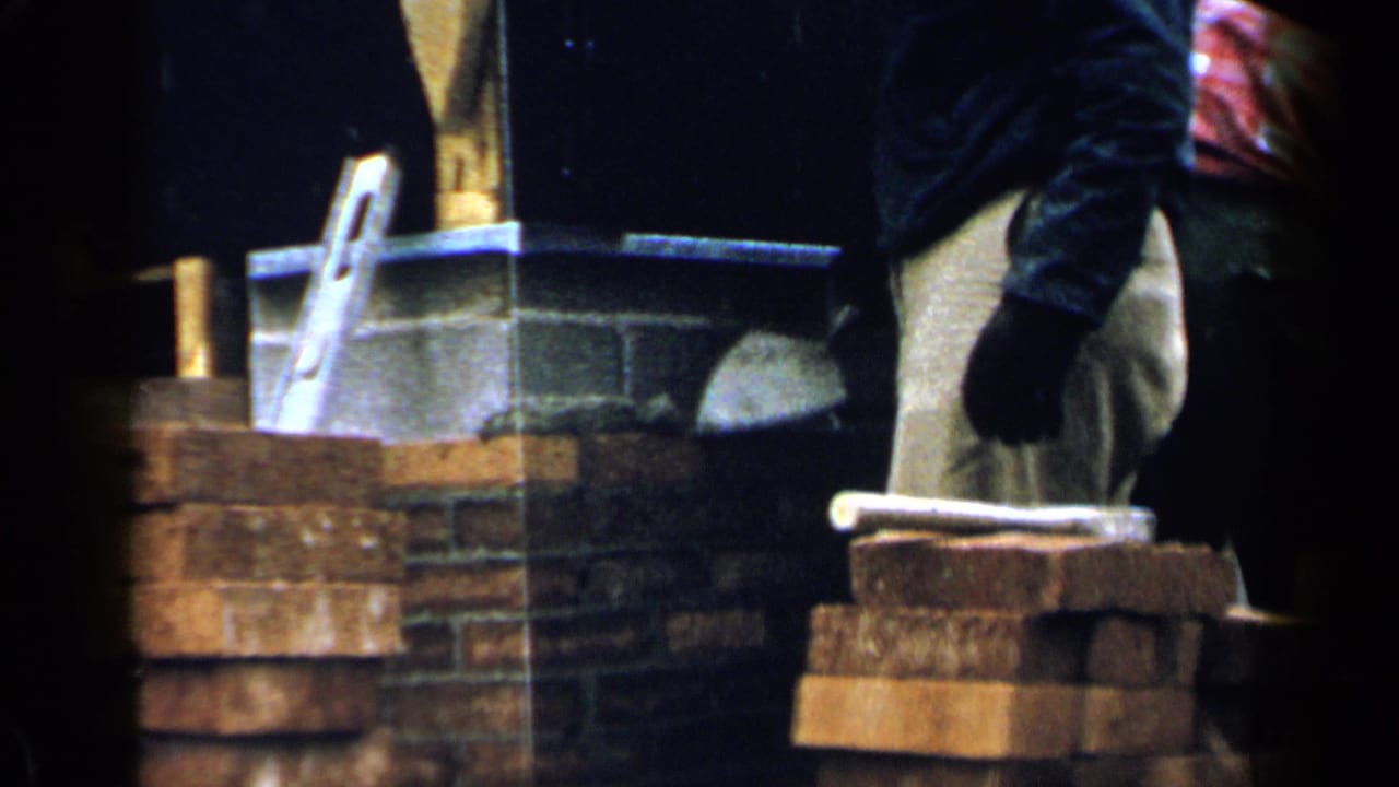 Worker in tan pants stands beside stacked bricks and a partially built chimney.
