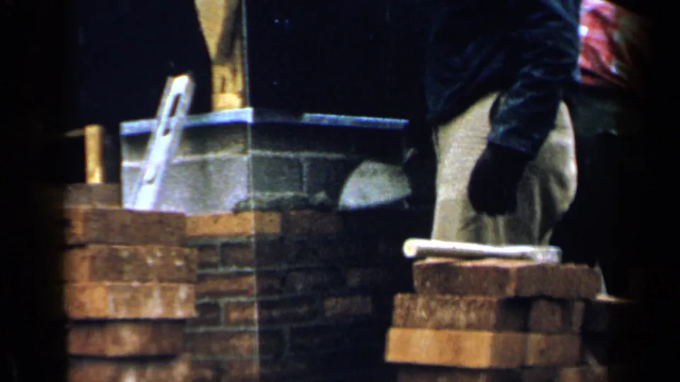 1957 Birmingham home-movie frame showing a worker in tan pants near brick stacks, mortar, and masonry tools.