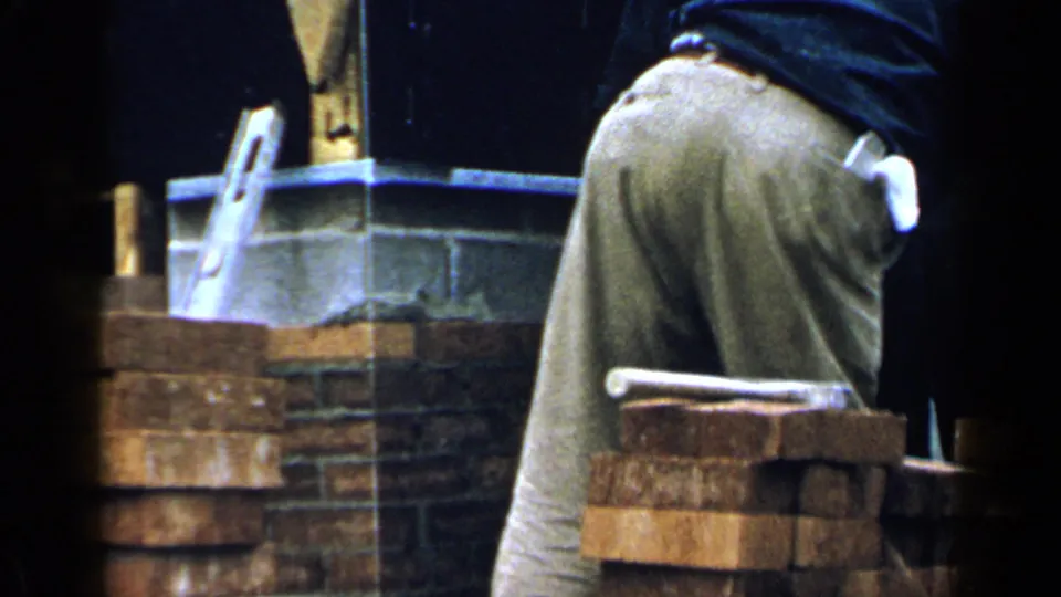 1957 Birmingham frame of a worker leaning over a brick chimney area with tools and fresh masonry visible.