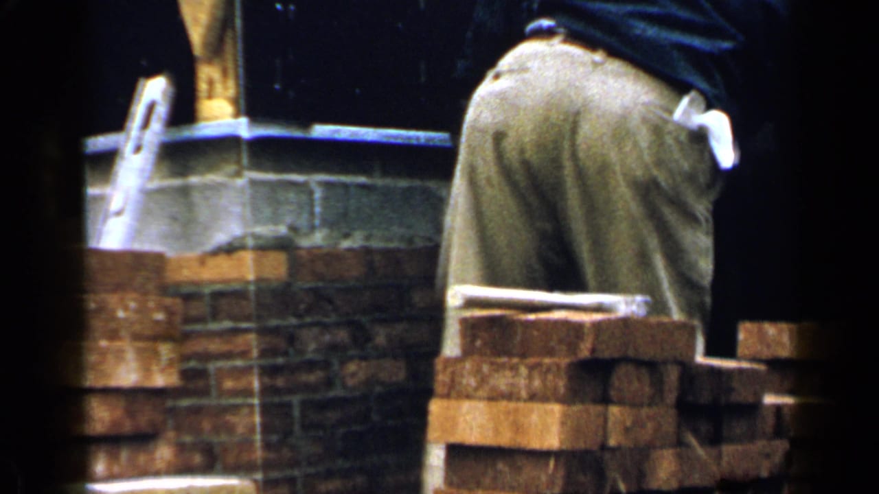 1957 Birmingham home-movie frame showing a worker from behind at a brick wall beside a house under construction.