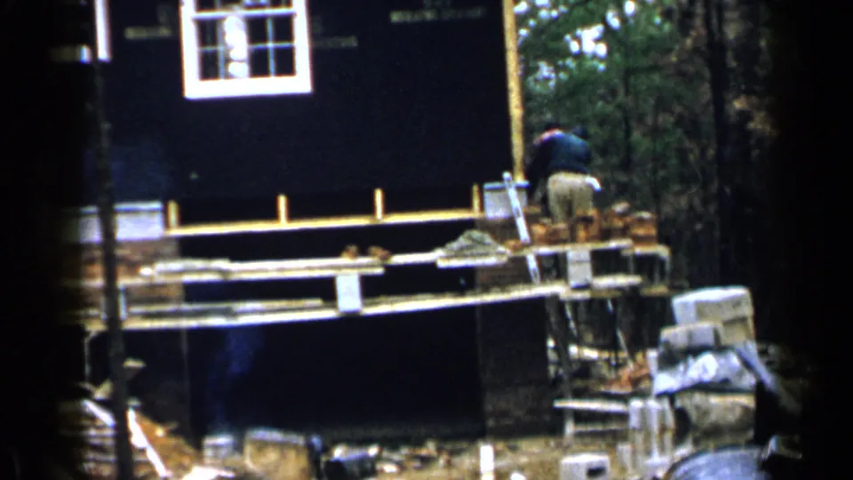 Wide 1957 Birmingham frame of a house under construction with a worker, brick stacks, framing, and trees.