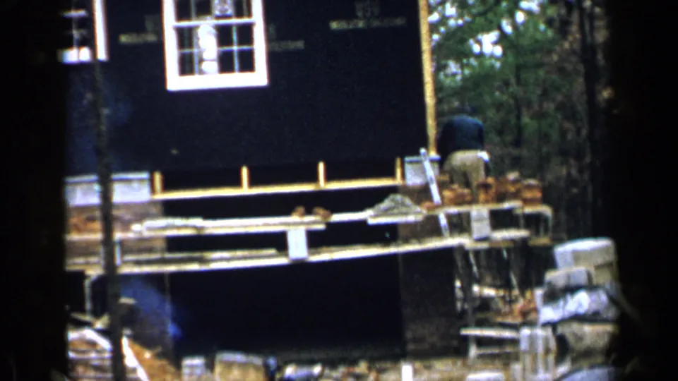 1957 Birmingham home-movie frame with a worker bending over bricks beside a partially built house and wooded background.