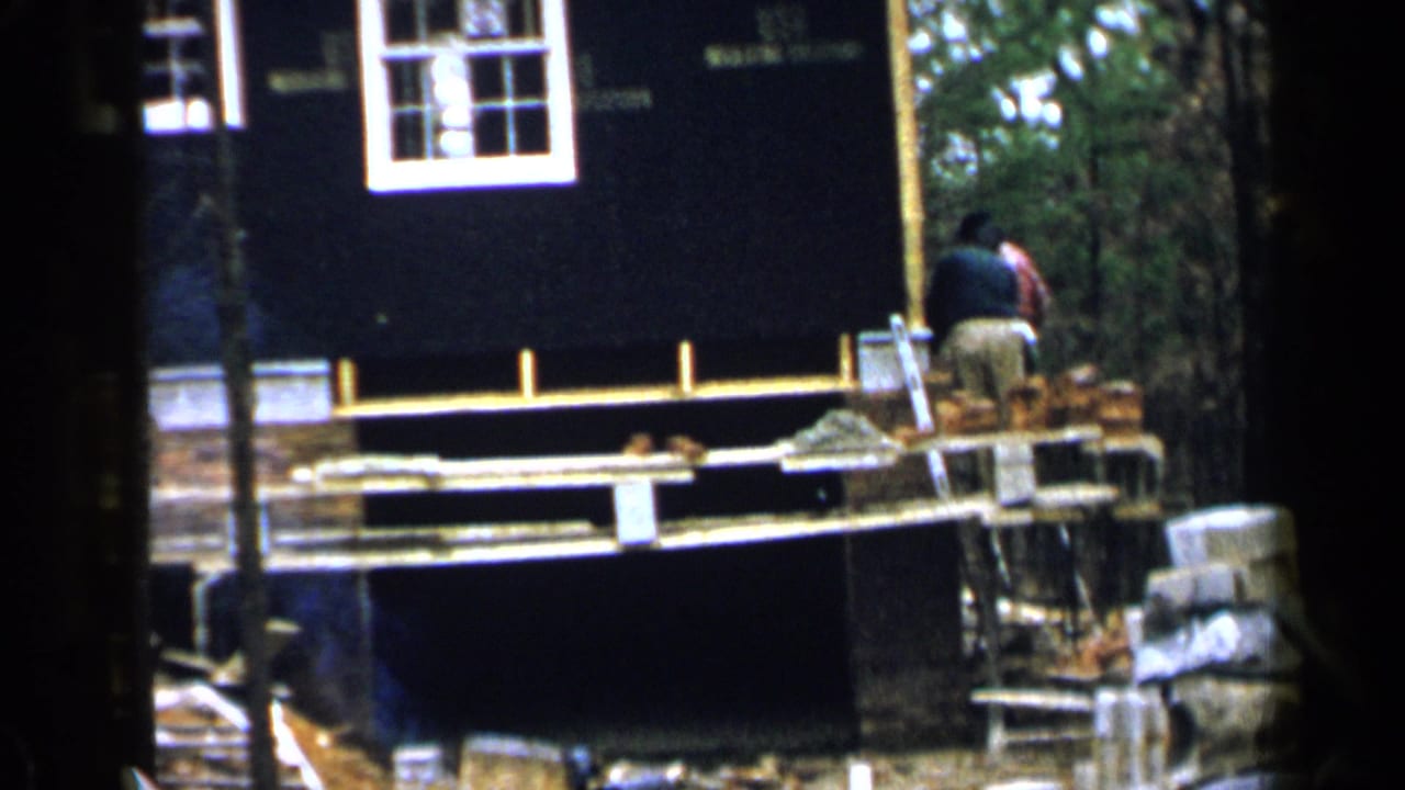 Man working on a wooden scaffold beside a dark house wall with a white-framed window.