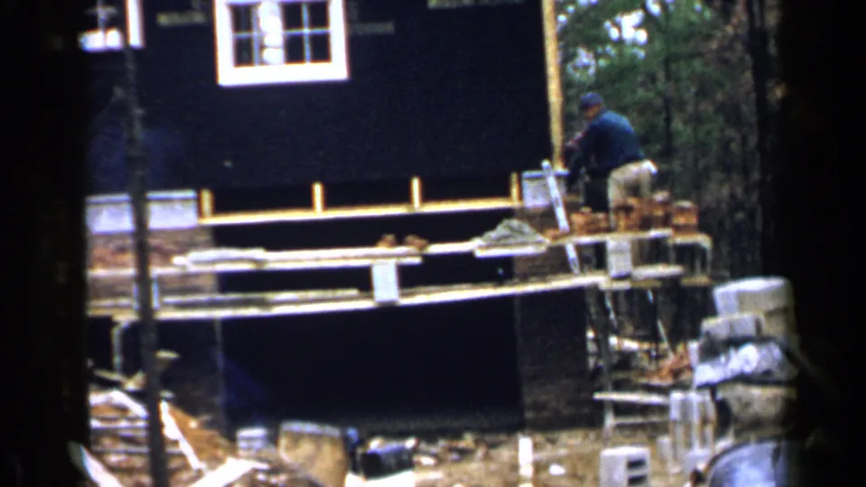 The bricklayer bends toward the scaffold work area, with stacked bricks and the house exterior filling the frame.