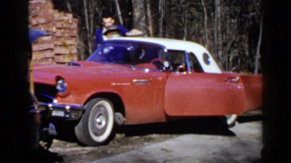 A home-movie frame shows the young man beside the red and white convertible, with the driveway and trees behind him.