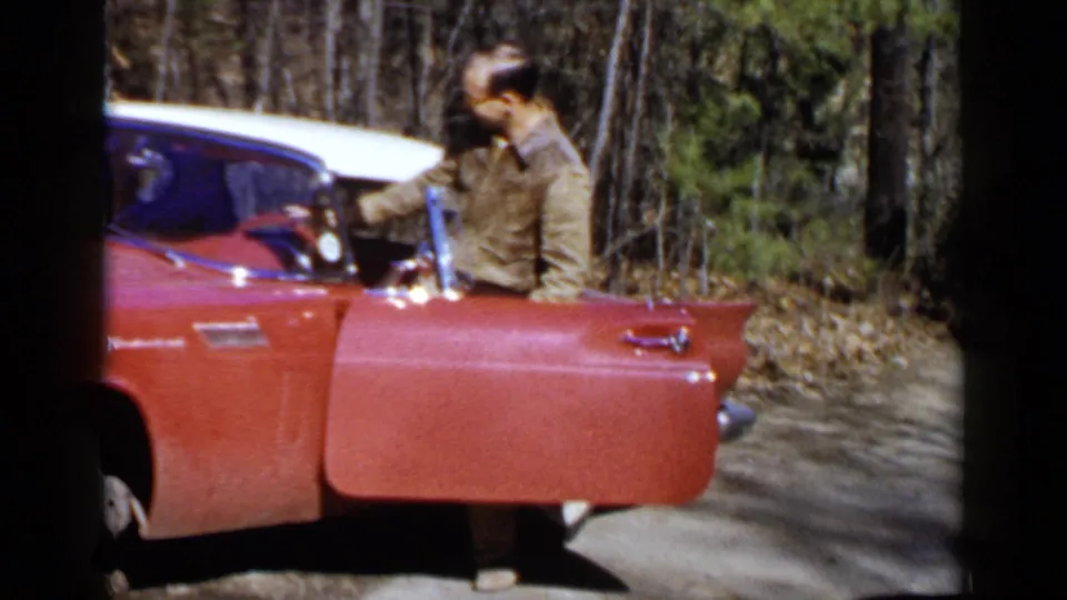 The man stands close to the red and white Thunderbird, leaning toward the driver's area on a wooded Birmingham roadside.