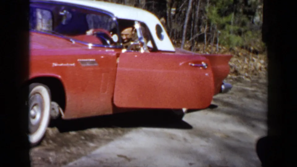 The man appears seated behind the wheel of the 1957 red and white Thunderbird, with the open door facing the wooded road.