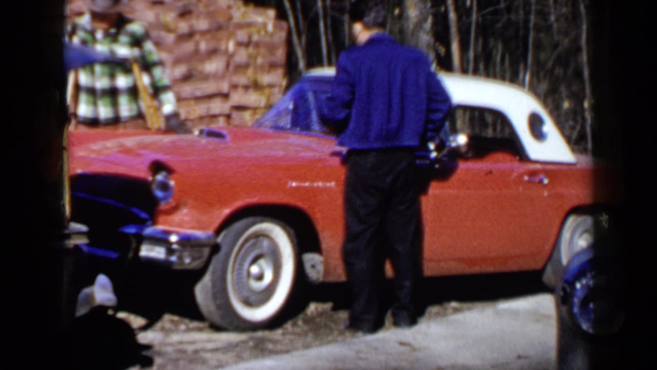Two men stand near a red car with a white roof beside a stack of bricks.