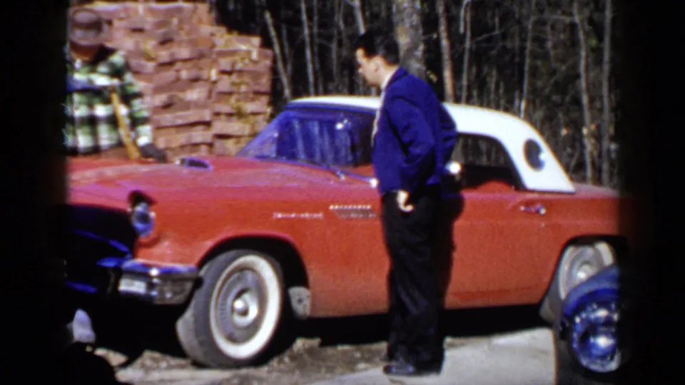 A man stands near the driver side of a red Ford Thunderbird-style car as another handles materials beside a brick stack.