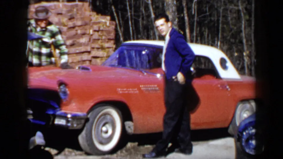 In the Birmingham 1957 footage, a man turns toward the camera beside the red car while another remains near stacked bricks.