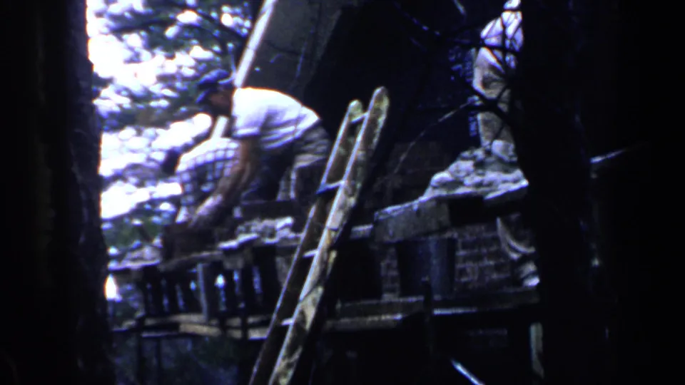 Dim home-movie frame showing a person bending near a ladder, wood or metal materials, and trees in an outdoor work area.