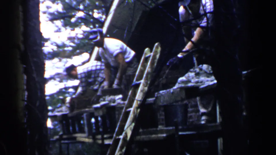 1957 Birmingham home-movie frame showing two adult men working on a raised wooden structure beside a ladder, viewed through dark tree trunks.