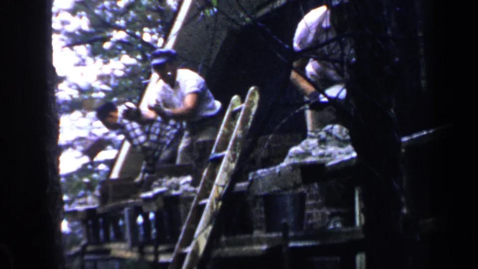 1957 Birmingham frame with a man in a white shirt and cap near a ladder as another man bends over boards on an outdoor structure.