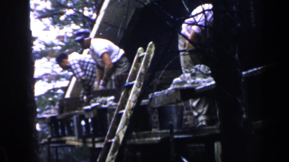 1957 Birmingham home-movie still of two men working on a rough wooden platform, with a ladder and surrounding trees framing the scene.