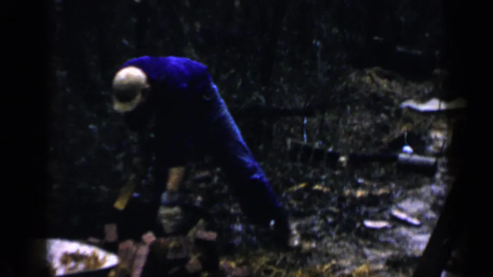 1957 Birmingham frame showing an adult man in dark blue bending over bricks or blocks on the ground in a wooded outdoor area.