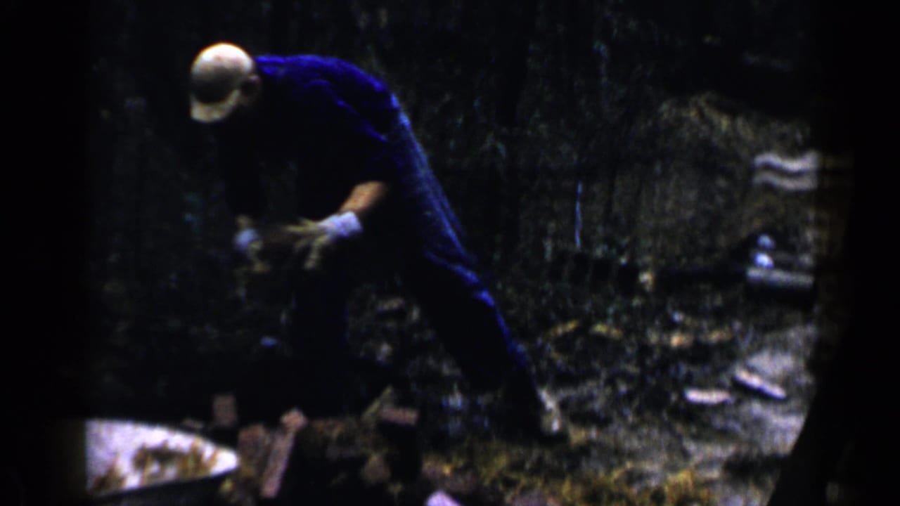 Man in a hard hat bends over bricks in a wooded outdoor work area.