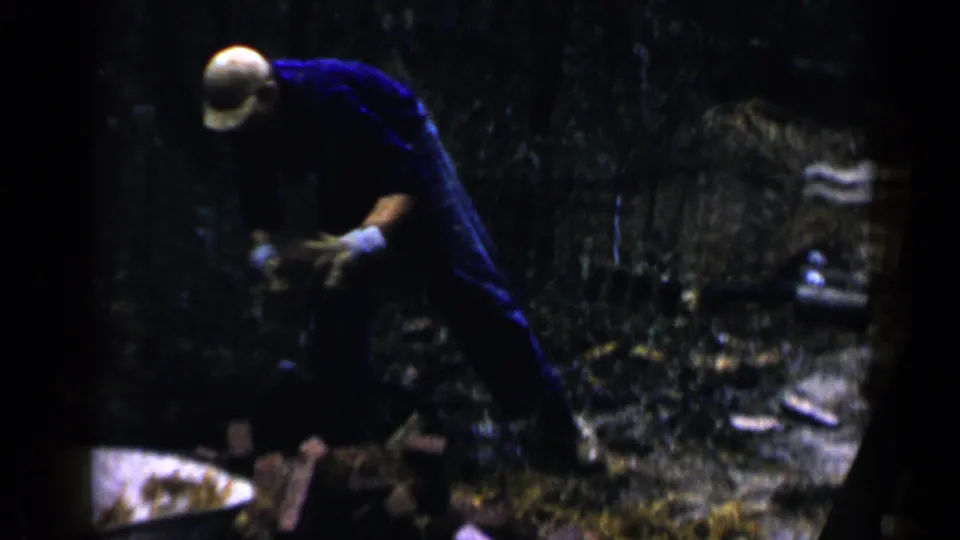 In 1957 Birmingham footage, a worker in a hard hat and blue shirt bends near bricks at a shaded outdoor construction site.