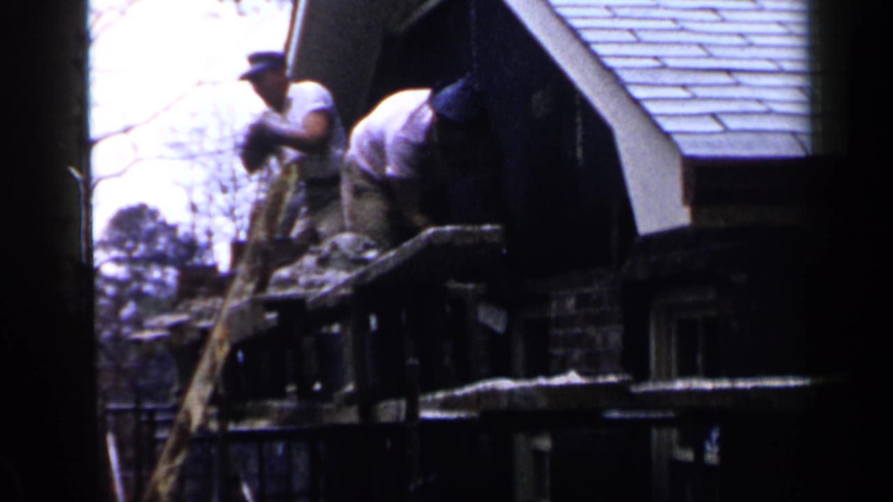 Two men work near a house roofline from a wooden scaffold.