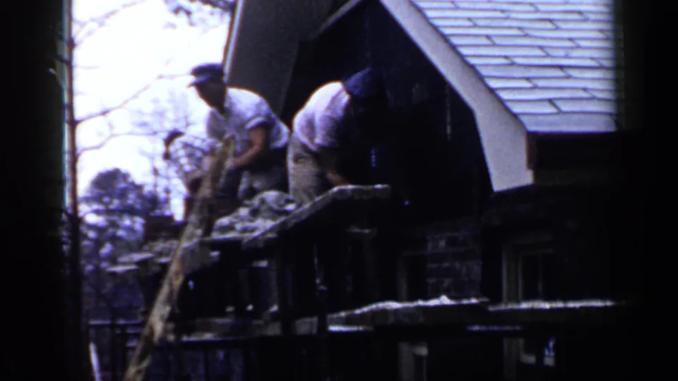 Workers in caps lean over scaffold planks by a shingled house roof in archival 1957 Birmingham footage.