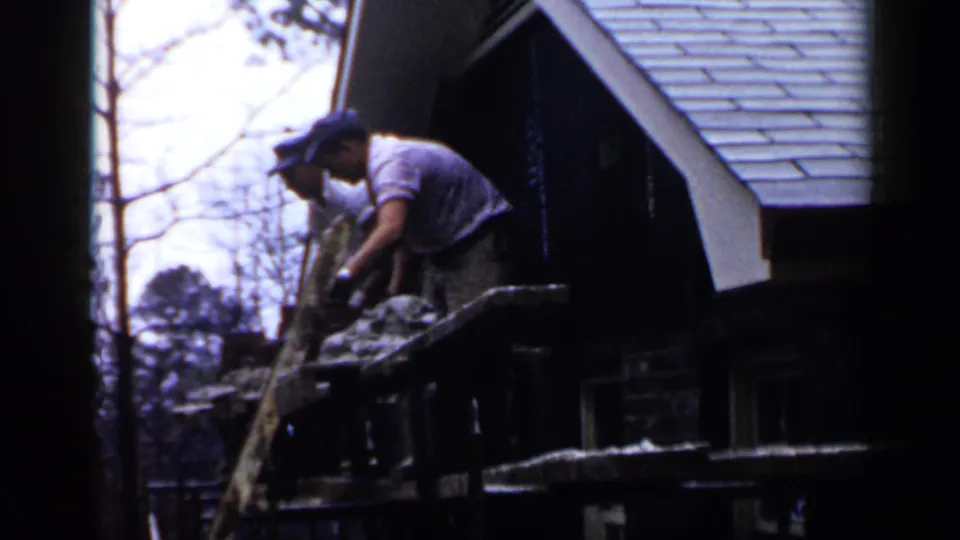 A man bends over a scaffold at the roofline of a Birmingham house, with another worker partly obscured nearby.
