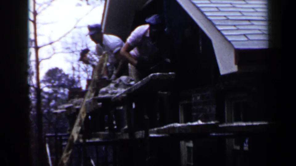 Two men stand on wooden scaffolding outside a brick house in 1957 Birmingham, Alabama, with roof shingles visible above.