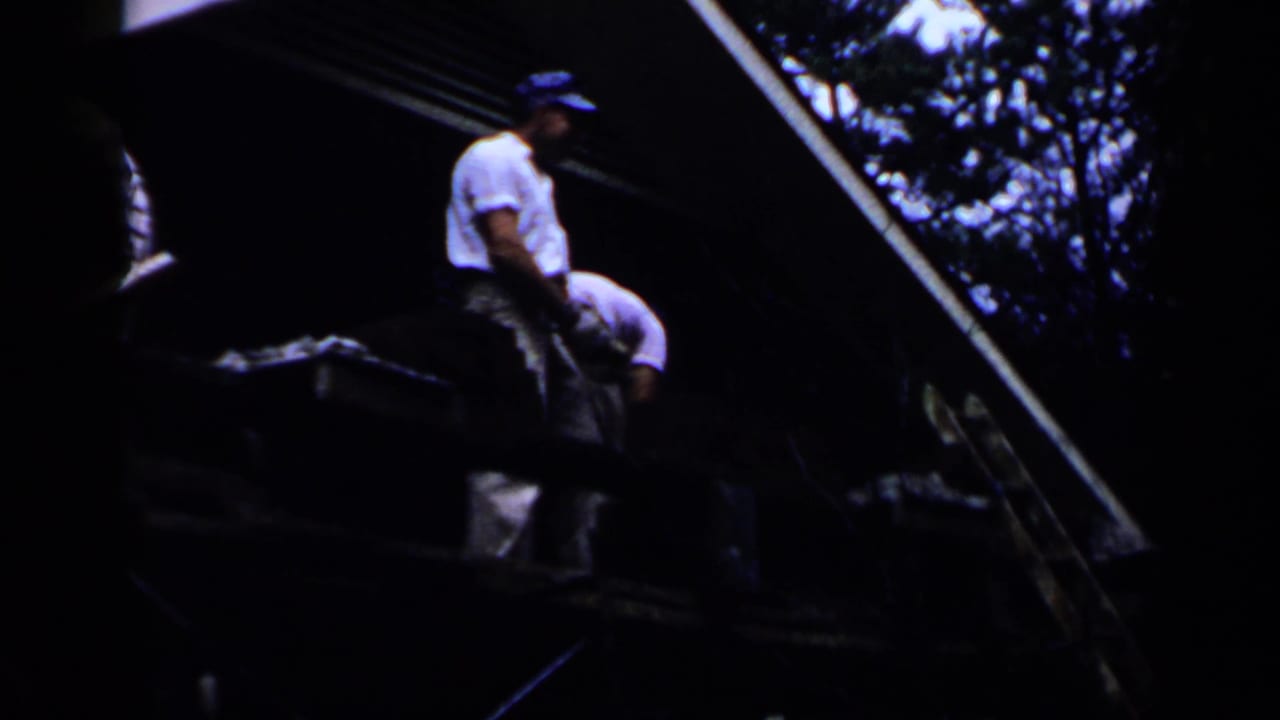In 1957 Birmingham home-movie footage, two adult men in white shirts and blue caps appear on a sloped roof or platform near trees.