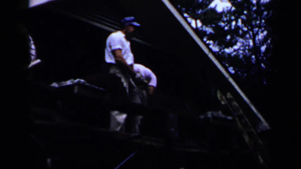 In 1957 Birmingham home-movie footage, two adult men in white shirts and blue caps appear on a sloped roof or platform near trees.