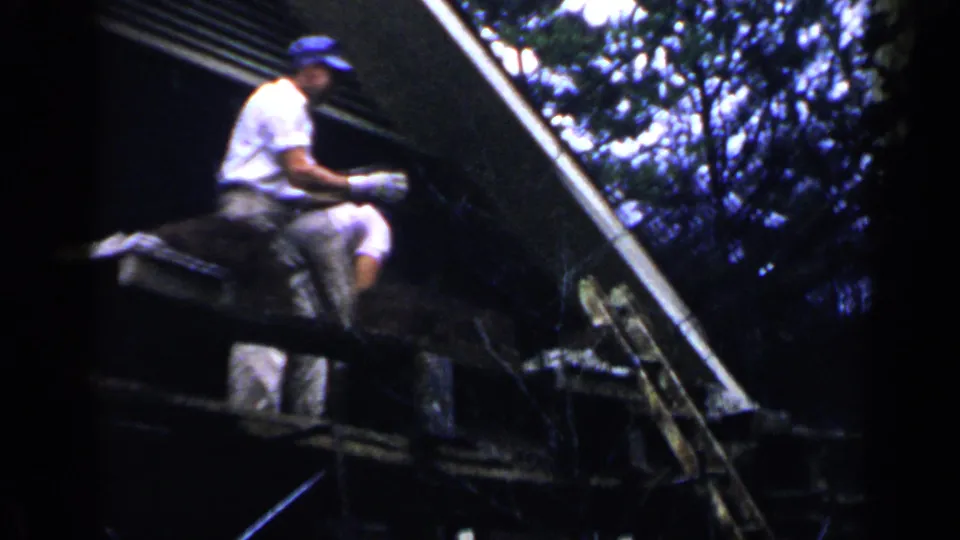 A man in a white shirt and blue cap works on a roof or raised platform in 1957 Birmingham, with trees and a roofline behind him.