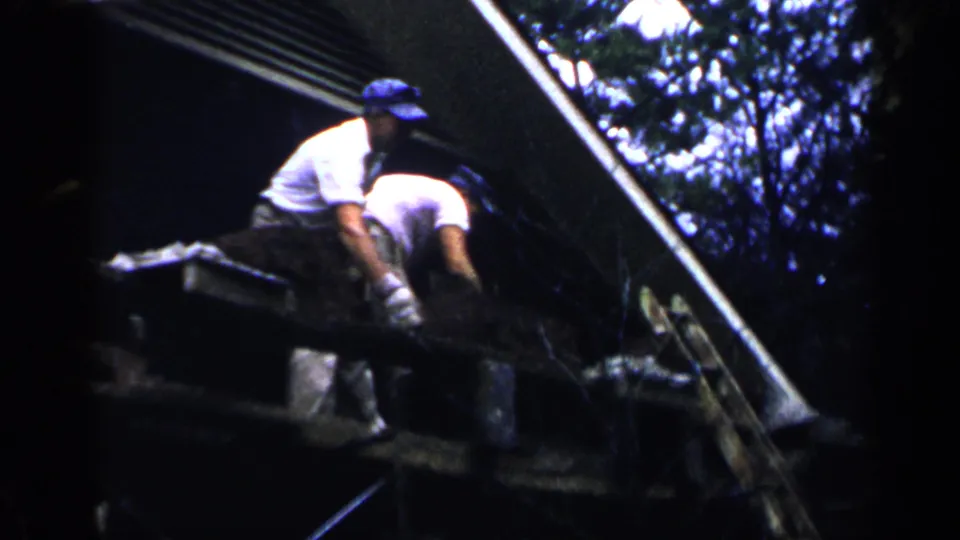 Two men in white shirts work close together on a sloped roof area in Birmingham, Alabama, in 1957, with a ladder and buckets nearby.